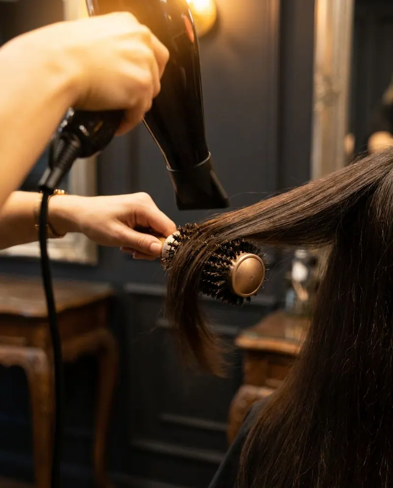 Friseurin föhnt mit Rundbürste langes dunkles Haar in einem Salon mit dunklen Wandpaneelen und warmem Licht
